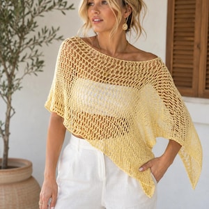 Puede incluir: Un poncho de ganchillo amarillo pálido con un patrón de rombos abierto y de punto suelto, con un amplio escote barco y un dobladillo asimétrico, usado sobre un top blanco sin tirantes y pantalones blancos.