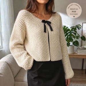 Puede incluir: Rebeca de crochet color crema con un lazo negro en el escote. La rebeca tiene un patrón de puntadas texturizadas y mangas largas. La imagen incluye el texto "CROCHET CARDIGAN DIGITAL PDF PATTERN".