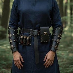May include: A dark blue long-sleeved tunic paired with black leather arm bracers featuring brass buckles. A black leather belt holds a rectangular pouch with a brass latch and a separate holder containing three small glass vials with cork stoppers.
