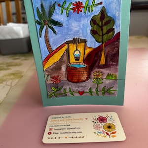 Op de afbeelding: Handgemaakte wenskaart met een kleurrijke landschapsillustratie met een put, palmboom en bloemen. De kaart is gemonteerd op een turquoise achtergrond. Een visitekaartje met bloemenillustraties en de tekst "Inspired by faith, hope & everyday beauty".