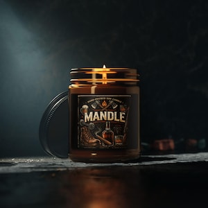 May include: A lit soy candle in a brown glass jar with a black lid. The label reads "MANDLE" with illustrations of a boot, axe, and whiskey. The candle is hand-poured.