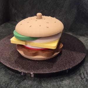 May include: A multi-layered toy hamburger constructed from stacked plastic components. The assembly features a tan bun top, a green lettuce layer, a yellow cheese slice, a white onion ring, a red tomato slice, and a brown meat patty base.