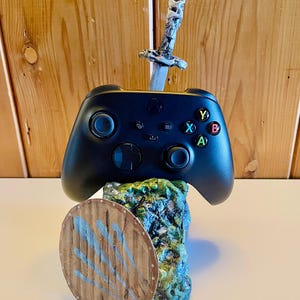 Puede incluir: Un mando de videojuegos negro sobre una base texturizada de color verde que imita una roca. Una pequeña espada plateada está incrustada en la parte superior de la base detrás del mando, y un escudo redondo de madera con marcas azules está fijado al frente.