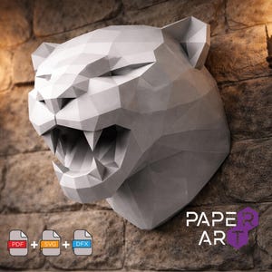Peut inclure: Une sculpture en papier géométrique low-poly représentant une tête de panthère rugissante en gris clair. Le design présente des facettes angulaires nettes et des dents visibles. Inclut des icônes de fichiers PDF, SVG et DFX, avec le texte PAPER ART.