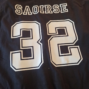 May include: A black t-shirt featuring the name SAOIRSE printed in white block letters above the large white number 32. The text and number are styled in a collegiate font with a thin black outline, centred on the back of the garment.