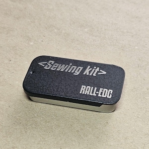 Könnte beinhalten: Kleine, rechteckige schwarze Nähbox mit dem Text "<Sewing kit>" und "RALL-EDC" auf dem Deckel. Der Kasten befindet sich vor einem neutralen, strukturierten Hintergrund.