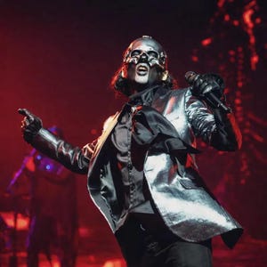 May include: A performer wearing a metallic silver blazer over a black button-down shirt with a black necktie. The performer has a skull-like mask with dark eye makeup and black gloves, holding a black microphone while standing under red stage lighting.