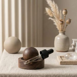 Puede incluir: Un difusor de aceites esenciales de madera con una base de madera marrón oscuro y una parte superior redonda de madera marrón oscuro. Un cuentagotas de vidrio con una tapa de goma negra descansa en la base. Una esfera beige y un jarrón con flores secas están en el fondo.