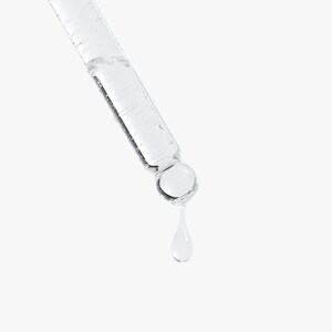 May include: A clear glass dropper with a liquid inside, angled diagonally against a white background. A single drop of liquid is suspended from the dropper's tip. The dropper is designed for dispensing liquids, likely for skincare or cosmetic products.