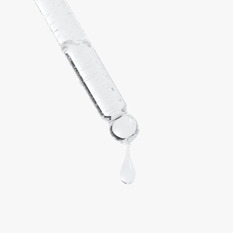 May include: A clear glass dropper with a liquid inside, angled diagonally against a white background. A single drop of liquid is suspended from the dropper's tip. The dropper is designed for dispensing liquids, likely for skincare or cosmetic products.