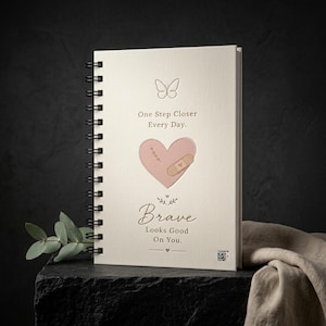 Op de afbeelding: Een spiraalgebonden notitieboek met een crèmekleurige kaft, voorzien van een lijntekening van een vlinder, een roze hart met een pleister en steken, en de tekst: One Step Closer Every Day. Brave Looks Good On You. Rechtsonder staat een kleine QR-code.