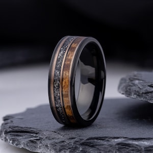May include: A black ring with a wood grain and stone inlay. The ring has a polished black band with a dark wood grain center and a band of stone-like material. The ring is displayed on a dark gray stone surface.