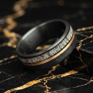 May include: A black ring with a band of white and brown material and a thin gold-colored stripe. The ring is set against a black marble surface.