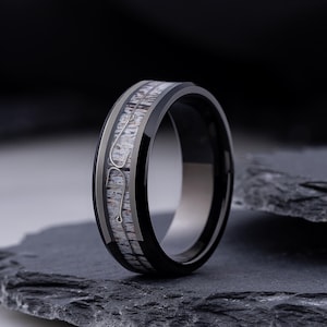 May include: A black band ring featuring a central inlay with a textured, light-colored organic pattern and two silver-toned fish hook silhouettes embedded within the inlay. The ring has a polished finish and a smooth, rounded outer profile.