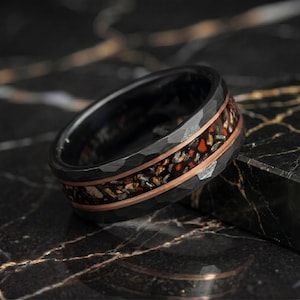 May include: A black hammered metal ring featuring a central inlay of crushed multi-colored stone fragments, including red, gold, and silver tones, bordered by two thin copper-colored bands that encircle the entire circumference of the ring.