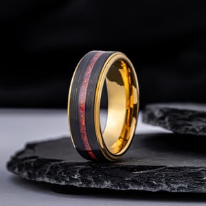 May include: A ring featuring a brushed black exterior with a central inlay of red crushed material. The interior of the band is polished gold, and the edges are finished with a gold-toned trim, creating a distinct contrast between the dark and metallic surfaces.