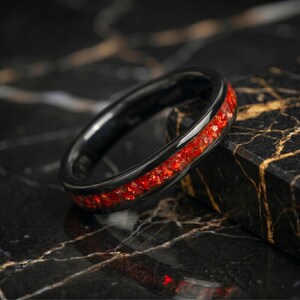 May include: A black ring featuring a central inlay of crushed red stone fragments. The band has a smooth, polished finish and a rounded profile, showcasing a vibrant contrast between the dark metal and the textured, sparkling red inlay.