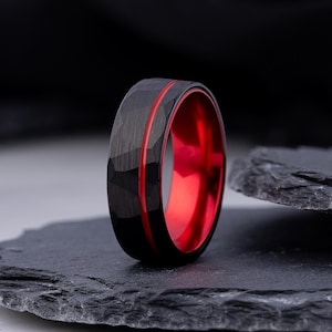 May include: A black ring with a hammered texture on the outer surface and a thin red groove running horizontally around the centre. The interior of the band is a vibrant, polished red, creating a distinct contrast with the dark exterior finish.