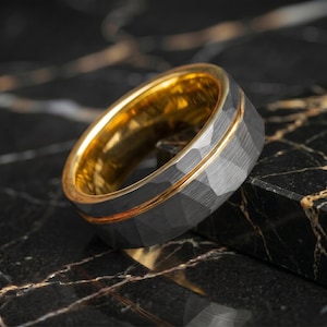 May include: A ring featuring a dark grey, faceted outer band with a brushed finish and a polished gold-tone inner band. A thin, recessed gold-tone groove separates the faceted exterior from the smooth, reflective interior surface of the ring.
