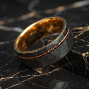May include: A dark gray hammered metal ring featuring a thin, copper-colored guitar string inlay running horizontally around the center of the band, with a smooth, polished gold-toned interior finish.