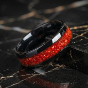 May include: A black band ring featuring a central inlay of crushed red material with a reflective, textured finish. The ring has a smooth, polished black exterior surface that contrasts with the vibrant, granular red inlay running around the center.