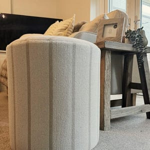May include: A round ottoman upholstered in light grey fabric with vertical stripes. The stripes are a slightly darker shade of grey. The ottoman is in a living room setting, near a wooden side table and a sofa.