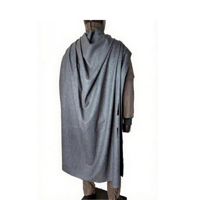 May include: A gray cape with a textured, speckled appearance. The cape drapes over a person's shoulders and back, reaching almost to the ankles. The person is wearing a brown jacket and pants. The cape has a button closure on the side.