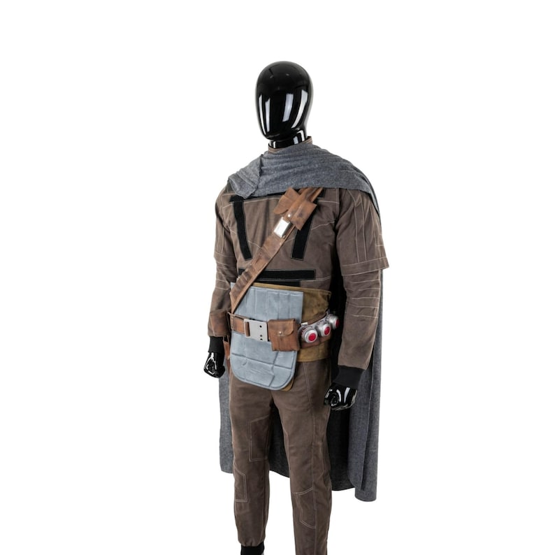 May include: A brown costume with a gray cape, displayed on a black mannequin. The costume includes a long-sleeved shirt, pants, and a utility belt with pouches and a red-lensed device. The mannequin is set against a white background.