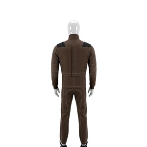 May include: A brown jumpsuit with white stitching and black shoulder accents. The jumpsuit has a high collar and long sleeves. The sleeves and shoulders have a geometric design. The pants are straight-legged.