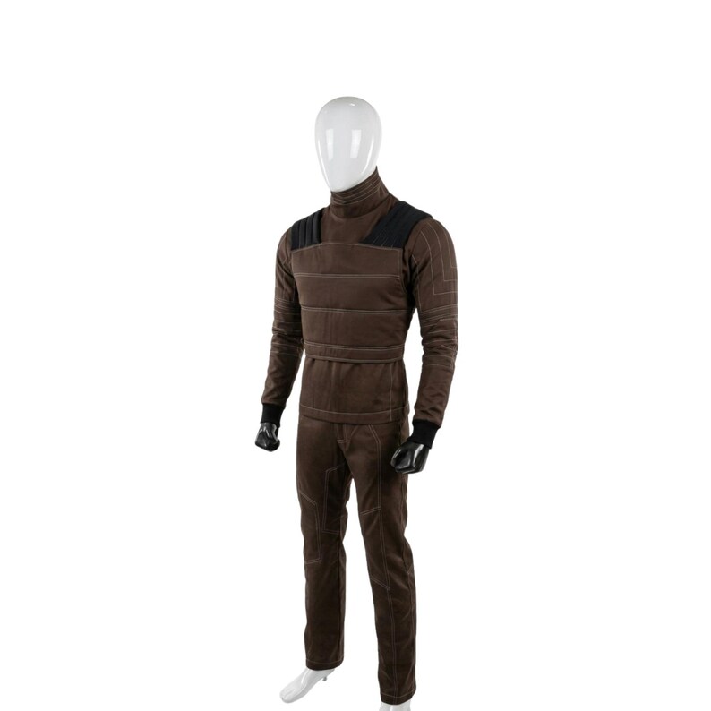 May include: A brown jumpsuit with a high collar, black shoulder accents, and white stitching. The long-sleeved top and pants are worn by a mannequin. Black gloves and white shoes complete the outfit.