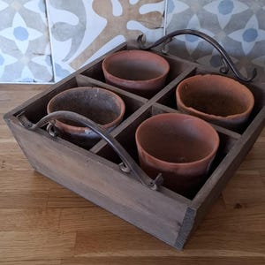 May include: A wooden tray containing four terracotta pots. The tray is divided into four compartments and features metal handles. The pots are reddish-brown and appear empty. The tray rests on a wooden surface.