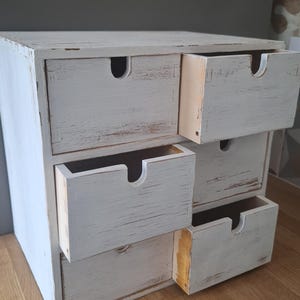 Puede incluir: Un pequeño mueble de cajones de madera pintado de blanco con seis cajones. El mueble tiene un acabado envejecido, con la veta de la madera visible y un aspecto desgastado. Los cajones tienen un recorte curvo para las manijas. El mueble está sobre una superficie de madera.
