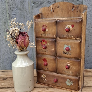 May include: A wooden wall-mounted storage cabinet with seven drawers, each featuring a red knob and hand-painted floral details. The drawers are labelled Needles, Buttons, Utility, Snaps, Pins, Thread, and Utility. Beside it sits a beige ceramic vase.