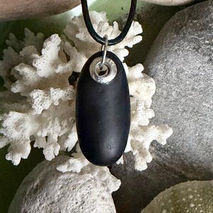 Peut inclure: Un collier avec un pendentif ovale noir et une bélière argentée. Le pendentif est suspendu à un cordon noir. Le fond présente du corail blanc et des pierres grises, créant une esthétique naturelle et balnéaire.