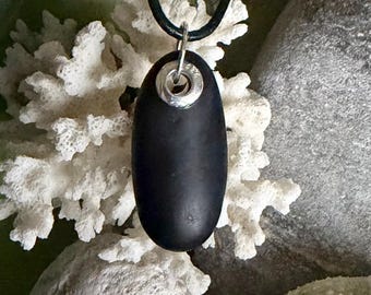 Può includere: Una collana con pendente ovale nero e anello argentato. Il pendente è appeso a un cordino nero. Lo sfondo presenta coralli bianchi e pietre grigie, creando un'estetica naturale e balneare.