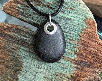 Può includere: Una collana con pendente in pietra nera con un anello in metallo color argento attaccato in alto. Il pendente è appeso a un cordino di cuoio nero. Il pendente è posto su uno sfondo di legno stagionato.