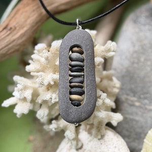 May include: A pendant featuring a large, oval-shaped gray stone with a hollow center. Inside the opening, a vertical stack of nine small, smooth, multi-colored pebbles is suspended on a thin wire. The pendant hangs from a simple black cord.