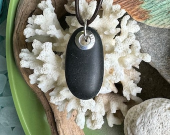 Può includere: Una collana con un ciondolo in pietra nera e un ciondolo in argento. Il ciondolo ha la forma di una pietra ovale liscia. La collana è su un cordoncino in pelle marrone.