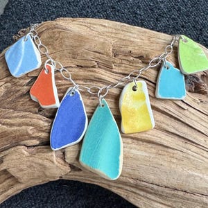 May include: A colorful necklace featuring irregularly shaped ceramic pendants in shades of blue, orange, yellow, and green, strung on a silver chain. The pendants have a textured appearance and are arranged in a cascading pattern. The necklace is displayed on a piece of driftwood.