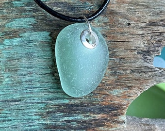 Può includere: Una collana con un ciondolo in vetro di mare verde chiaro, una ghiera in argento e un cordoncino in pelle nera. Il ciondolo è su una superficie in legno rustico.