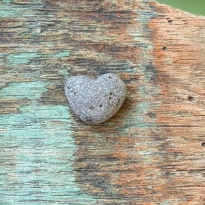 May include: A gray, heart-shaped stone with small dark spots rests on a weathered wooden surface. The wood has a mix of brown, teal, and natural wood tones, creating a rustic aesthetic. The stone is smooth and natural.