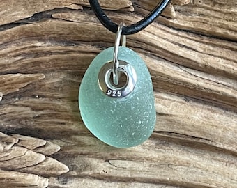 Può includere: Una collana con un ciondolo in vetro di mare verde chiaro, un anello d'argento e il numero 925 impresso su di esso. La collana è su un cordoncino di cuoio nero.