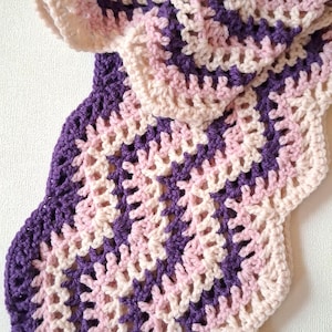 May include: A crocheted scarf featuring a wavy, chevron-like pattern in alternating stripes of deep purple, soft pink, and cream yarn. The ends of the scarf are finished with a fringe made of alternating light pink and cream yarn tassels.