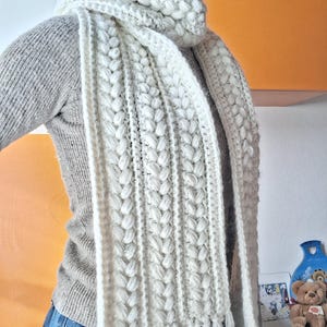 May include: A thick, off-white crocheted scarf featuring a textured cable-knit pattern with vertical braided columns. The scarf is wrapped around the neck and hangs down with long, straight fringe tassels at the bottom edges of the fabric.