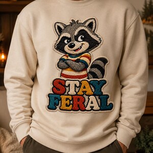 Peut inclure: Sweat-shirt crème avec un dessin de raton laveur souriant, les bras croisés, portant une chemise rayée. Le texte "STAY FERAL" est brodé en dessous en lettres colorées. Le sweat-shirt a un col rond et des manches longues.