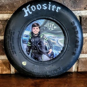 Op de afbeelding: Een zwarte Hoosier-raceband met een ronde inzet in het midden met een illustratie van een jonge autocoureur in een zwart pak die een helm vasthoudt, met raceauto's en een geblokte vlag op de achtergrond. De zijkant van de band toont 3.20/4.5-5.