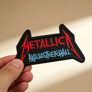 May include: Embroidered black patch featuring the word "METALLICA" in red and "...AND JUSTICE FOR ALL" in blue. The patch has a unique arrow-like shape with red lightning bolt accents. A hand is holding the patch.