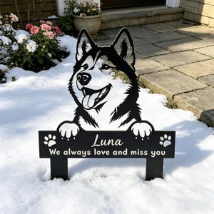 Puede incluir: Letrero de jardín de metal negro con un diseño de perro husky sonriente. El letrero incluye el nombre "Luna" y el texto "We always love and miss you". Huellas de patas en el letrero. El letrero está en la nieve.