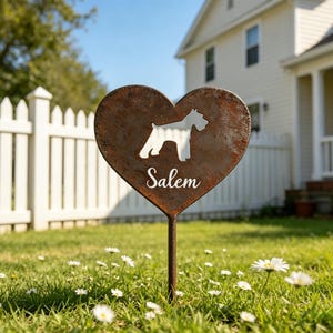 May include: A heart-shaped, rusty metal garden stake featuring a dog silhouette and the name "Salem". The stake is set in a grassy garden with daisies, a white picket fence, and a house in the background.