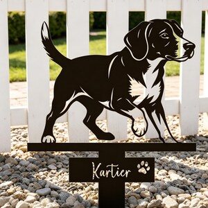 Puede incluir: Estaca de jardín de metal negro con silueta de perro, con el nombre "Kartier" y una huella de pata. El perro se muestra caminando, contra una valla blanca. La estaca está colocada en un lecho de pequeñas piedras.